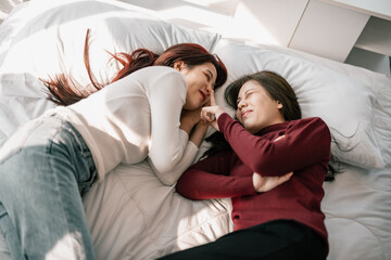 Fototapeta na wymiar Asian lesbian partners smile, relax, laugh, affectionately pinch and kiss each other's cheeks while lying on the bed. A combination of personal charm, a good sense of humor, and a romantic moment.