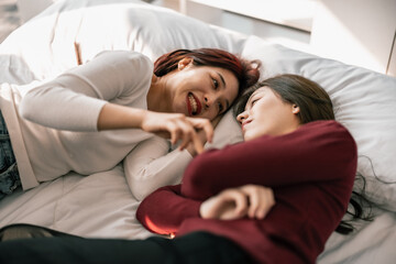 Asian lesbian partners smile, relax, laugh, affectionately pinch and kiss each other's cheeks while lying on the bed. A combination of personal charm, a good sense of humor, and a romantic moment.