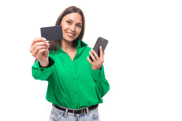 smiling well-groomed slim european young brunette woman with makeup in a green shirt shows a bank card with a mockup and a smartphone on a white background with copy space for a banner