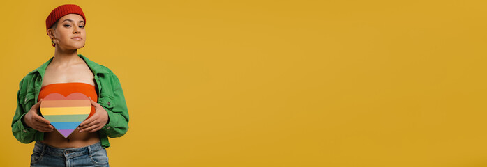 Confident young woman holding heart shaped rainbow flag against yellow background