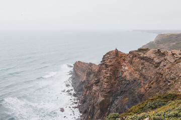 Adventurous man standing on the edge of a cliff enjoys the view of the Atlantic coast in the Odemira region of southwestern Portugal. Wandering the Rota Vicentina