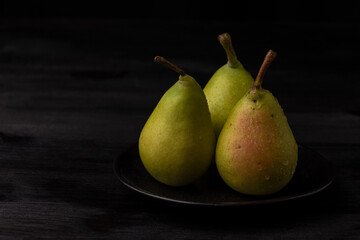 Close-up of three green pears with water drops in black plate on dark wooden table, horizontal, with copy space