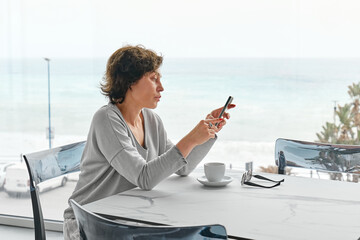 Worried woman check social networks from phone while drinking coffee alone in cafe in the morning near panoramic seaside window.