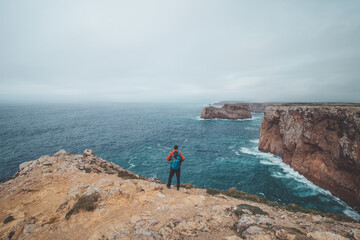 Fototapeta na wymiar Black-haired adventurer stands at the end of Cape Cabo de Sao Vicente in the southwest of Portugal in the Algarve region. Man is enjoying his freedom. Wandering of Fisherman Trail