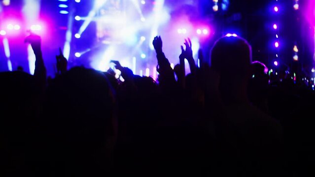 People jump and dance at the evening concert with light show.Fans raise their hands in front bright colorful strobe lights on stage. Crowd is dancing, filming performance rock band, throwing party.
