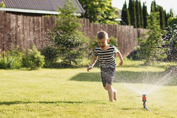 A little wet boy runs barefoot on the lawn next to the sprinkler. The concept of a happy, carefree childhood and vacation - obrazy, fototapety, plakaty