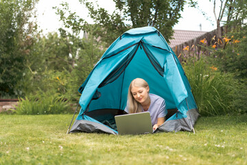 A young girl lying on the grass in the tourist tent with a laptop and communicates through a video call.