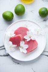 Plate with raw fresh tuna steaks, flowers and limes, vertical shot on a white marble background, high angle view