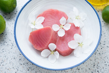 Obraz na płótnie Canvas Blue and white plate with fresh uncooked tuna steaks and flowers, horizontal shot on a light-grey granite background, middle closeup