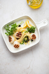 Grilled sea scallops with spaghetti, lemon zest and parsley in a beige serving pan, high angle view on a light-beige stone background, vertical shot