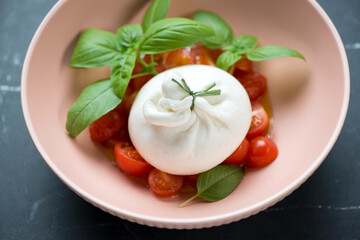 Burrata served with cherry tomatoes and green basil in a roseate bowl, middle closeup on a black marble background, selective focus
