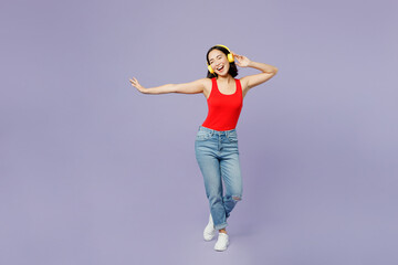 Full body fun young woman of Asian ethnicity she wear casual clothes red tank shirt headohones listen to music dance isolated on plain pastel light purple background studio portrait Lifestyle concept.