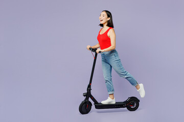 Full body side view young woman of Asian ethnicity she wear casual clothes red tank shirt riding electric scooter isolated on plain pastel light purple background studio portrait. Lifestyle concept.