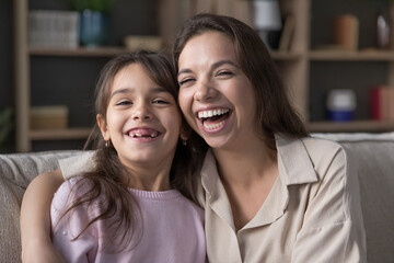 Cheerful pretty young mom hugging cute preschool kid girl looking at camera with toothy gaps smile, laughing, enjoying motherhood, home leisure. Parent and child head shot portrait