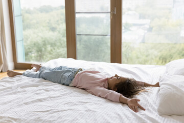 Positive cute little girl kid resting on double bed in bedroom, lying on back on soft mattress, stretching body, enjoying leisure, laziness, relaxation on white linen, blanket, laughing, having fun