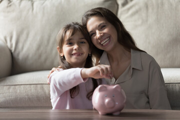 Obraz na płótnie Canvas Happy young mom and kid girl saving money for education, dropping coins into piggy bank, caring for finance, economy, learning investment, looking at camera, smiling. Home family portrait
