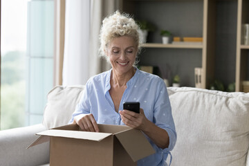Happy blonde mature customer woman opening cardboard paper box, holding mobile phone, using express...