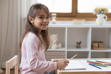 Home portrait of happy adorable little preschool kid girl drawing in paper album, sitting at small desk in playroom, using colorful pencils, looking at camera with toothy gap smile