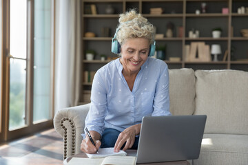 Cheerful older retired woman in wireless big earphones studying at home, writing notes at computer, watching webinar, lecture, looking at laptop, smiling, using Internet technology for education
