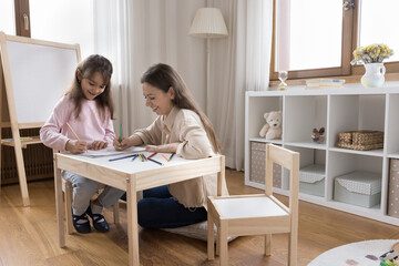 Happy preschool little child girl and positive mom drawing in colorful pencils in kids playroom,...