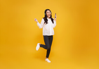 Happy Asian girl listening favourite music playlist on moblie phone application with wireless headphones and dancing isolated on yellow studio background.