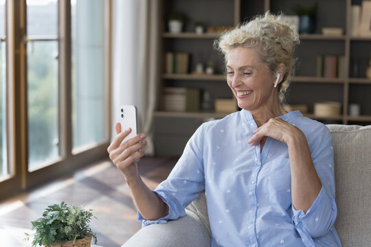 Cheerful elder mature woman talking on video conference call at home, using small wireless earphone in ear, holding smartphone, enjoying smiling, laughing, enjoying online conversation