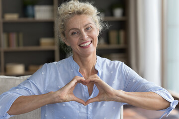 Cheerful positive pretty mature lady applying finger heart shaped hands to chest, looking at...