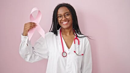 African american woman doctor holding breast cancer awareness pink ribbon over isolated pink background - Powered by Adobe