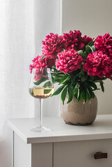 A glass of white wine and a bouquet of red peonies in a ceramic vase on a white wooden chest of drawers