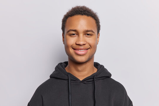 Portrait of handsome dark skinned adult man feels cheerful smiles pleasantly dressed in casual black sweatshirt looks directly at camera isolated over white background. Happy emotions concept
