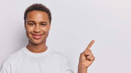 Cheerful dark skinned man promots product that demands discovery smiles pleasantly points index finger on copy space gives advice or advertises product dressed in casual t shirt isolated on white wall