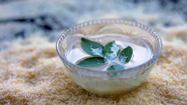 Oryza sativa and Oryza glaberrima or raw lachkari kolam rice grains on a black surface along with a glass bowl containing rice water in it with some rose leaves on top, used in beauty products and fac