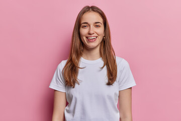 Fototapeta premium Portrait of lovely European millennial girl with straight hair smiles toothily being in good mood dressed in casual white t shirt isolated over pink studio background. Positive human emotions