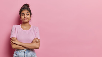 Horizontal shot of young Indian girl with hair bun keesps arms folded stands in thoughtful pose concentrated aside tries to make decision dressed in casual t shirt and jeans pink background.