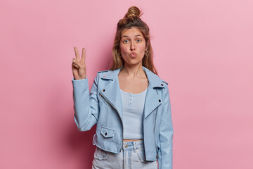 Lovely dark haired woman with hair bun keeps lips folded makes peace gesture expresses positive emotions and joy dressed in blue jacket and jeans isolated over pink background. Body language concept