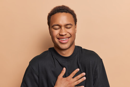 Photo of overjoyed dark skinned man keeps hand on chest laughs happily keeps eyes closed dressed in casual black t shirt isolated over brown background being in good mood. People and emotions concept