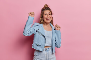 Studio shot of pretty cheerful young European woman radiating joy as she dances and has fun epresses enthusiasm and cheerfulness dressed in blue jacket and jeans isolated over pink background
