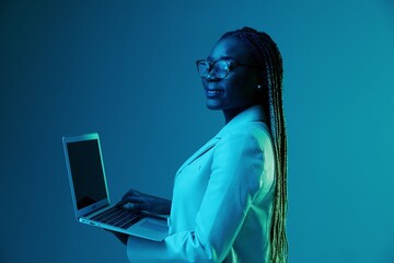 African African woman with laptop in glasses on a blue background in neon light, color mixed light. Data storage cyber security, hacker, big data.
