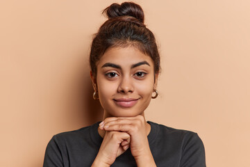 Portrait of charming Indian woman with dark hair combed in bun keeps hands under chin wears casual black t shirt and earrings looks directly at camera isolated over beige background. Studio shot