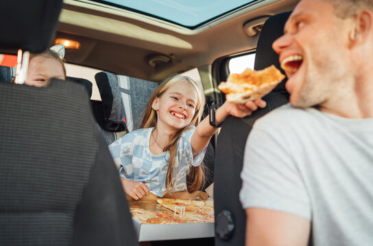 Portrait of positive smiling girl putting pizza piece in father's mouth while she sitting in child car seat . Happy childhood, fast-food eating or auto jorney lunch break concept image