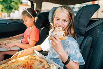 Portrait of positive smiling girl eating just cooked italian pizza sitting with a sister on car back seat in child car seats. Happy childhood, fastfood eating or auto jorney lunch break concept image.