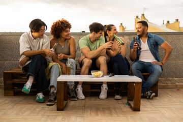 A group of multiracial young people are sitting on a rooftop of a flat in the open air. The 5 millennials are chatting while drinking beers. Concept of young people celebrating success, end of exams.