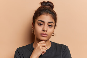 Headshot of lovely Indian woman keeps hand under chin focused directly at camera has calm expression dressed in casual black t shirt isolated over brown background. Human facial expressions.