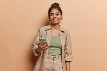 Studio shot of cheerful Indian girl with hair bun uses mobile phone for making video call or broadcasting livestream wears shirt and trousers isolated over brown background looks at device screen - 609666678