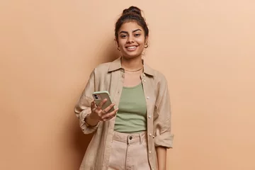 Studio shot of cheerful Indian girl with hair bun uses mobile phone for making video call or broadcasting livestream wears shirt and trousers isolated over brown background looks at device screen © WHstudio Leushin N