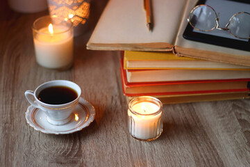 Cup of tea or coffee, plate of cookies, books, e-reader, pencil and lit candles on the table. Selective focus.