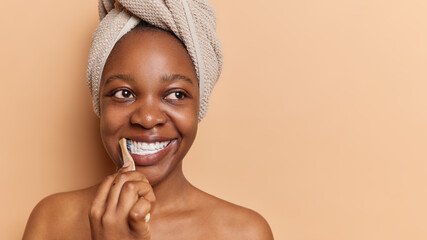 Horizontal shot of dark skinned woman with bath towel wrapped around her head demonstrates morning routine by attentively brushing teeth with toothbrush and toothpaste isolated over brown background