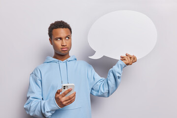 Pensive dark skinned adult man looks seriously at blank communication bubble holds mobile phone...