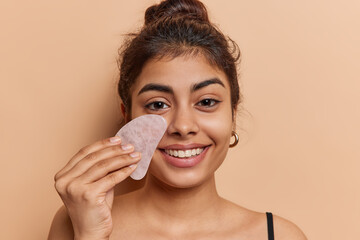 Close up shot of dark haired European woman massages face with guasha stone undergoes skin care procedures smiles toothily isolated over brown background. Facial massaging and rejuvenation concept