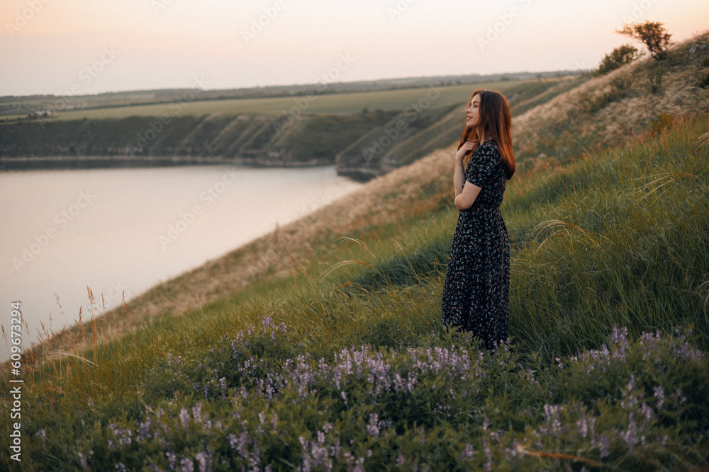 Wall mural Portrait of a beautiful young brunette woman in a summer dress at sunset among beautiful nature. The girl fixes her hair and looks into the distance. Place for text.